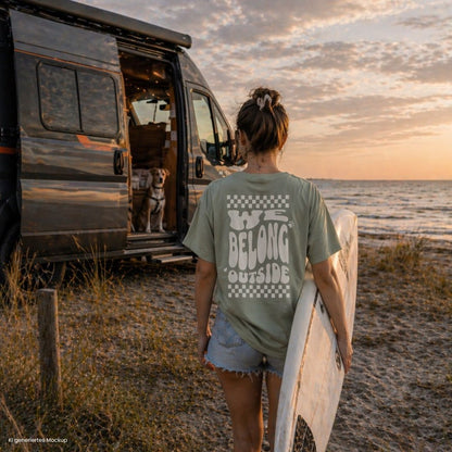 Person läuft am Strand mit Surfboard neben Camper und Hund und trägt salbeigrünes „we belong outside“ T Shirt in Farbe Bay, ideal für Vanlife und Camping